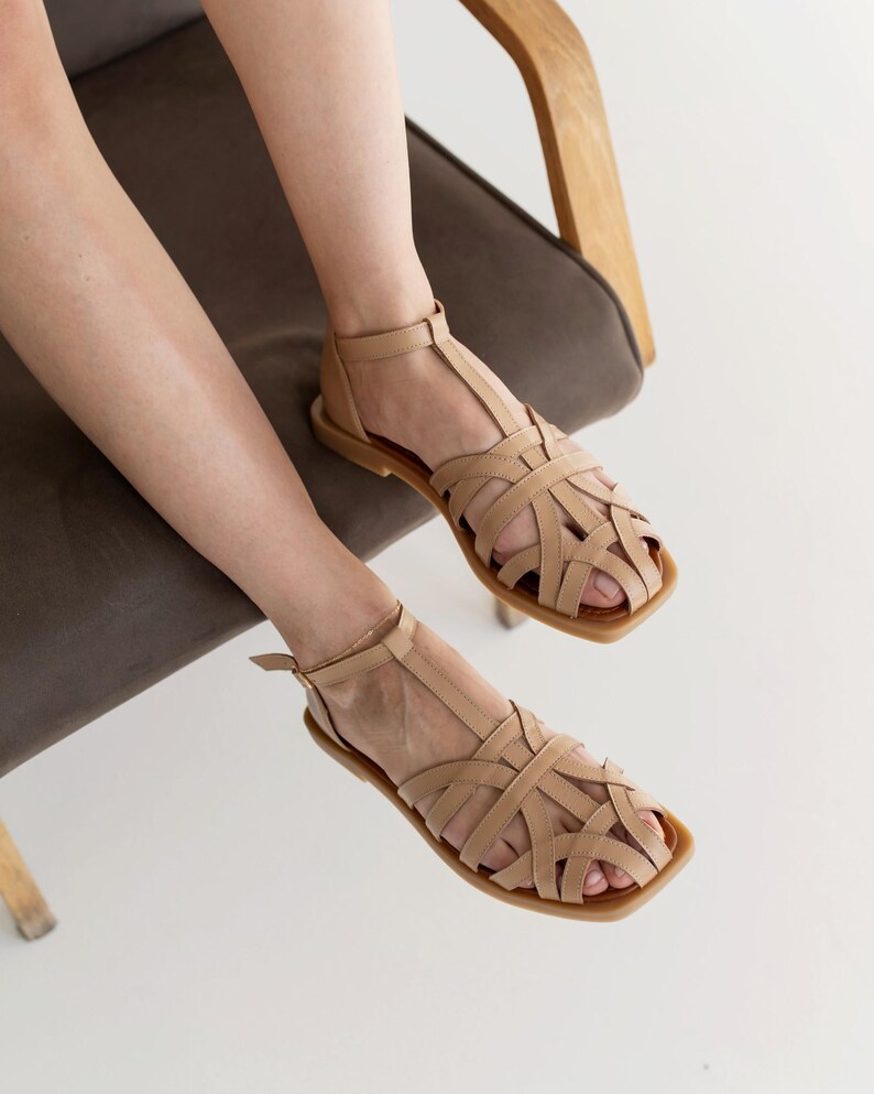 May include: Tan leather sandals with a woven design. The sandals have a square toe and a strap that goes around the ankle. The sandals are sitting on a brown chair.