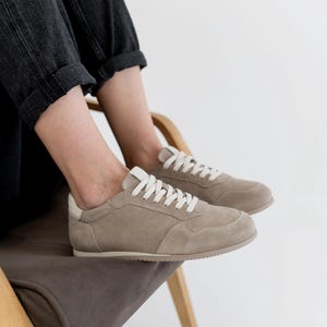 Pu&ograve; includere: Un paio di sneakers in camoscio beige con lacci bianchi e un rinforzo bianco sul tallone. Una persona con jeans grigio scuro &egrave; seduta su una sedia di legno con rivestimento grigio scuro, indossando le sneakers.