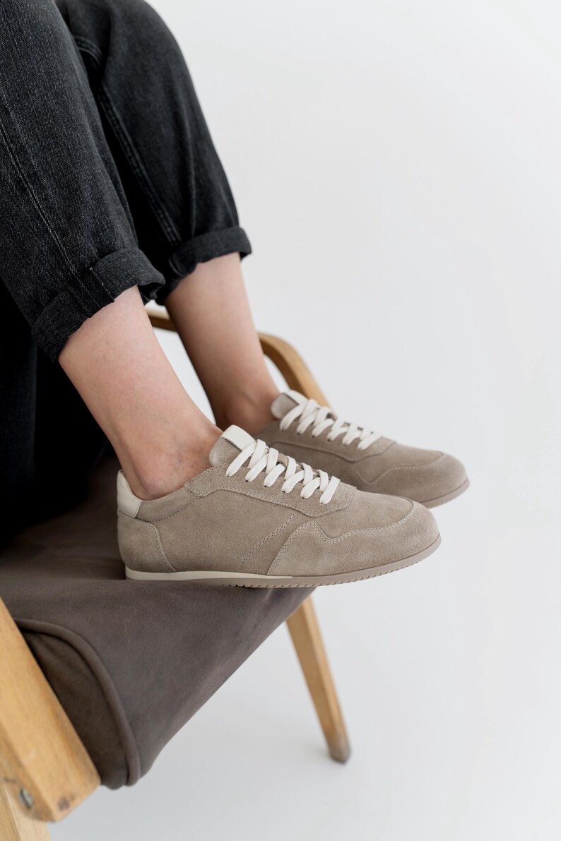 Pu&ograve; includere: Un paio di sneakers in camoscio beige con lacci bianchi e un rinforzo bianco sul tallone. Una persona con jeans grigio scuro &egrave; seduta su una sedia di legno con rivestimento grigio scuro, indossando le sneakers.