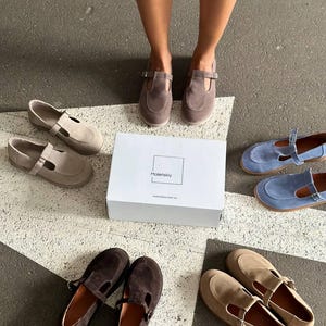 May include: An assortment of suede Mary Jane style shoes in various colors, including brown, tan, and blue, are displayed around a white box. The shoes have a strap and buckle closure. The box has the brand name "Maletskiy" printed on it.