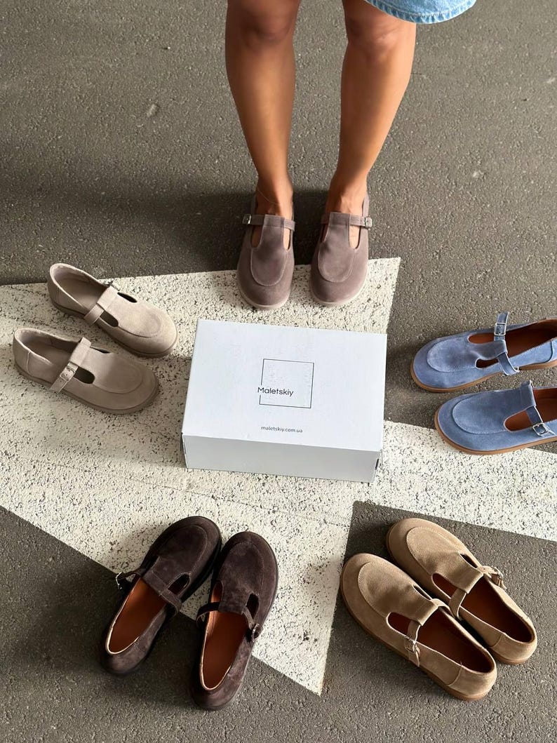May include: An assortment of suede Mary Jane style shoes in various colors, including brown, tan, and blue, are displayed around a white box. The shoes have a strap and buckle closure. The box has the brand name "Maletskiy" printed on it.