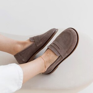 May include: A pair of taupe suede loafers with a classic penny loafer design. The shoes have a rounded toe and a low heel. A gold ankle bracelet is visible. The shoes are on a cream-colored surface.