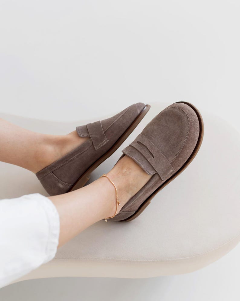 May include: A pair of taupe suede loafers with a classic penny loafer design. The shoes have a rounded toe and a low heel. A gold ankle bracelet is visible. The shoes are on a cream-colored surface.