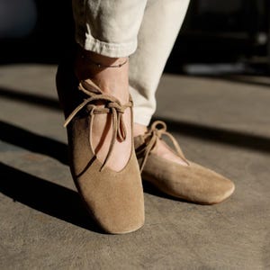 Side view of a woman wearing sand-colored suede Mary Jane flats with laces and beige trousers.