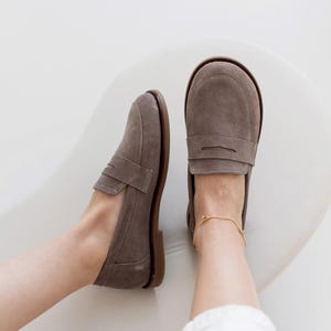 May include: A pair of gray suede loafers with a classic penny strap design. The shoes have a brown sole and are shown from a top-down perspective. A gold anklet is visible on the right ankle. The shoes are on a white surface.
