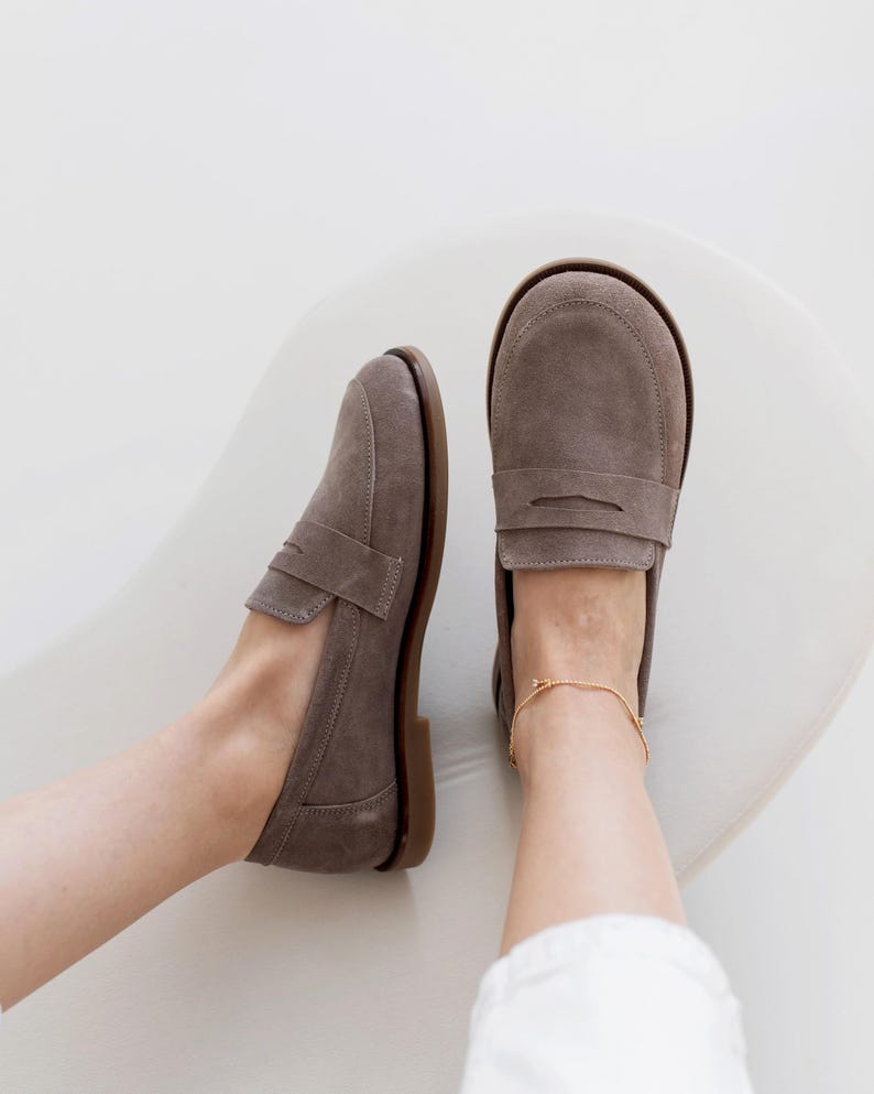 May include: A pair of gray suede loafers with a classic penny strap design. The shoes have a brown sole and are shown from a top-down perspective. A gold anklet is visible on the right ankle. The shoes are on a white surface.