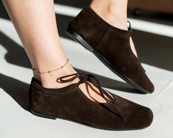 May include: Dark brown suede flats with a tie closure. The shoes have a rounded toe and a low heel. The image shows the shoes on a light surface, with one foot in the foreground and the other in the background.