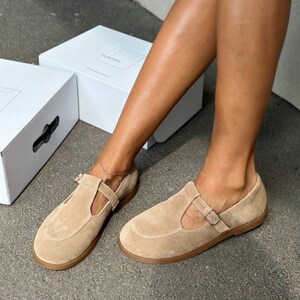 May include: Tan suede T-strap flats with a buckle closure. The shoes have a rounded toe and a flat sole. A white box is in the background.