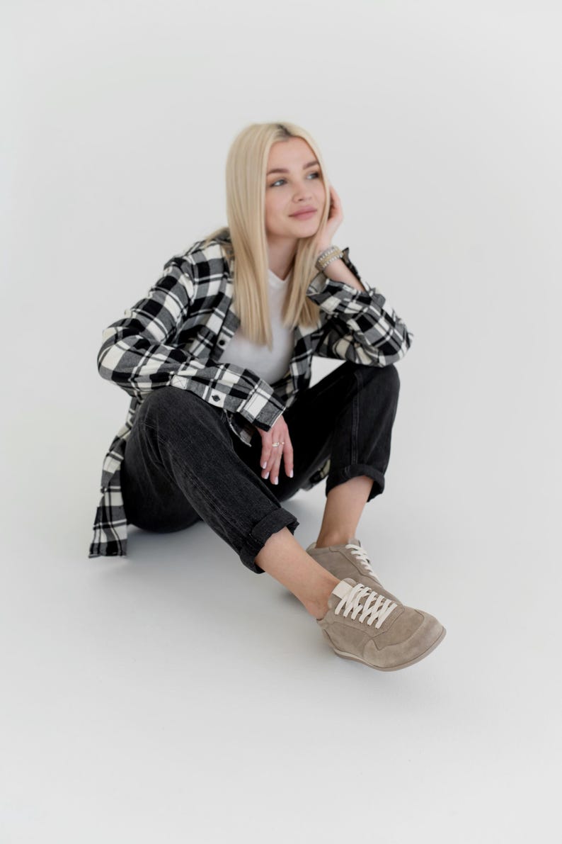 Pu&ograve; includere: Una persona indossa una maglietta bianca, jeans neri e una camicia a quadri bianchi e neri. La persona indossa anche scarpe da ginnastica beige con lacci bianchi. L'outfit &egrave; casual e comodo.