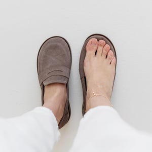 May include: A pair of taupe suede loafers, one worn on a foot with a gold anklet. The other shoe is empty. The shoes have a classic penny loafer design with a rounded toe and a low heel. The background is white.