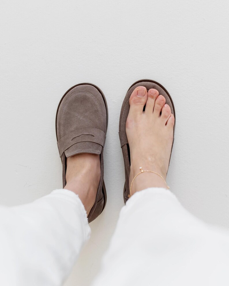 May include: A pair of taupe suede loafers, one worn on a foot with a gold anklet. The other shoe is empty. The shoes have a classic penny loafer design with a rounded toe and a low heel. The background is white.