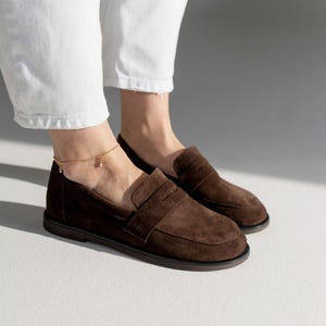 May include: A pair of dark brown suede loafers featuring a classic penny slot design across the vamp, a rounded toe, and a flat dark sole. The shoes are worn with white trousers and a delicate gold chain anklet with a small dangling charm.