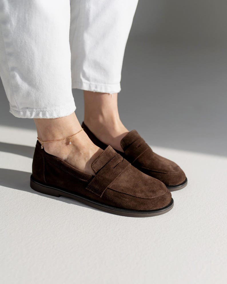 May include: A pair of dark brown suede loafers featuring a classic penny slot design across the vamp, a rounded toe, and a flat dark sole. The shoes are worn with white trousers and a delicate gold chain anklet with a small dangling charm.