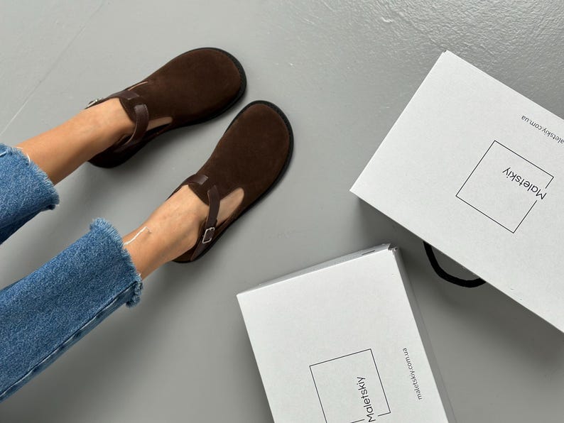May include: A pair of brown suede shoes with a T-strap design. The shoes are dark brown and have a buckle closure. Two white boxes with the brand name "Maletskiy" are also in the image.