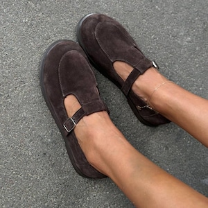 May include: A pair of brown suede Mary Jane style shoes. The shoes have a rounded toe, a strap across the top with a buckle, and a small cutout on the side. The shoes are on a gray surface.