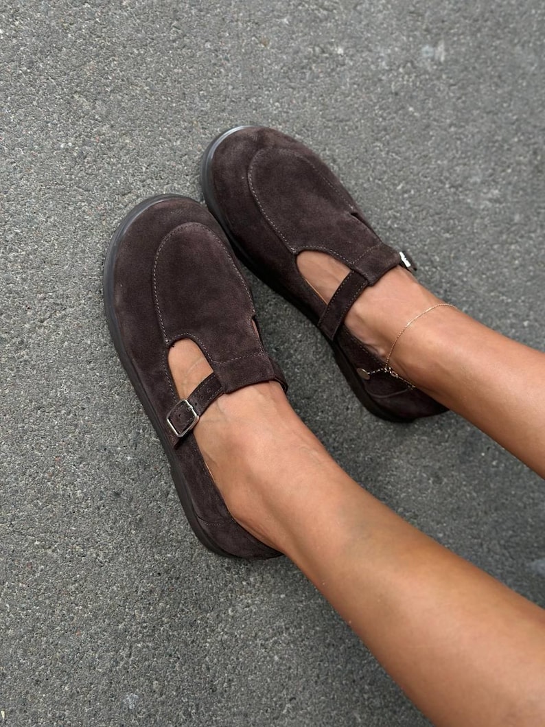 May include: A pair of brown suede Mary Jane style shoes. The shoes have a rounded toe, a strap across the top with a buckle, and a small cutout on the side. The shoes are on a gray surface.