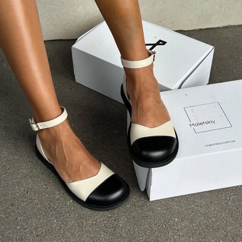 Women's barefoot shoes: beige and black leather anatomical Mary Janes with a wide toe box, a delicate ankle strap, and a minimalist zero-drop sole, handmade in Ukraine. These elegant cap toe flats are showcased in a minimalist boutique setting on a concrete plinth. Keywords: barefoot shoes, anatomical Mary Janes, wide toe box, two tone flats, beige and black shoes, minimalist flats, zero drop shoes, handmade in Ukraine, quiet luxury.