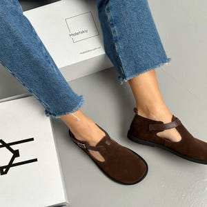 May include: Brown suede flats with a T-strap design and buckle closure. The shoes have a rounded toe and a dark brown leather strap. The shoes are displayed with a white box that has the text "Maletskiy" and "maletskiy.com.ua".