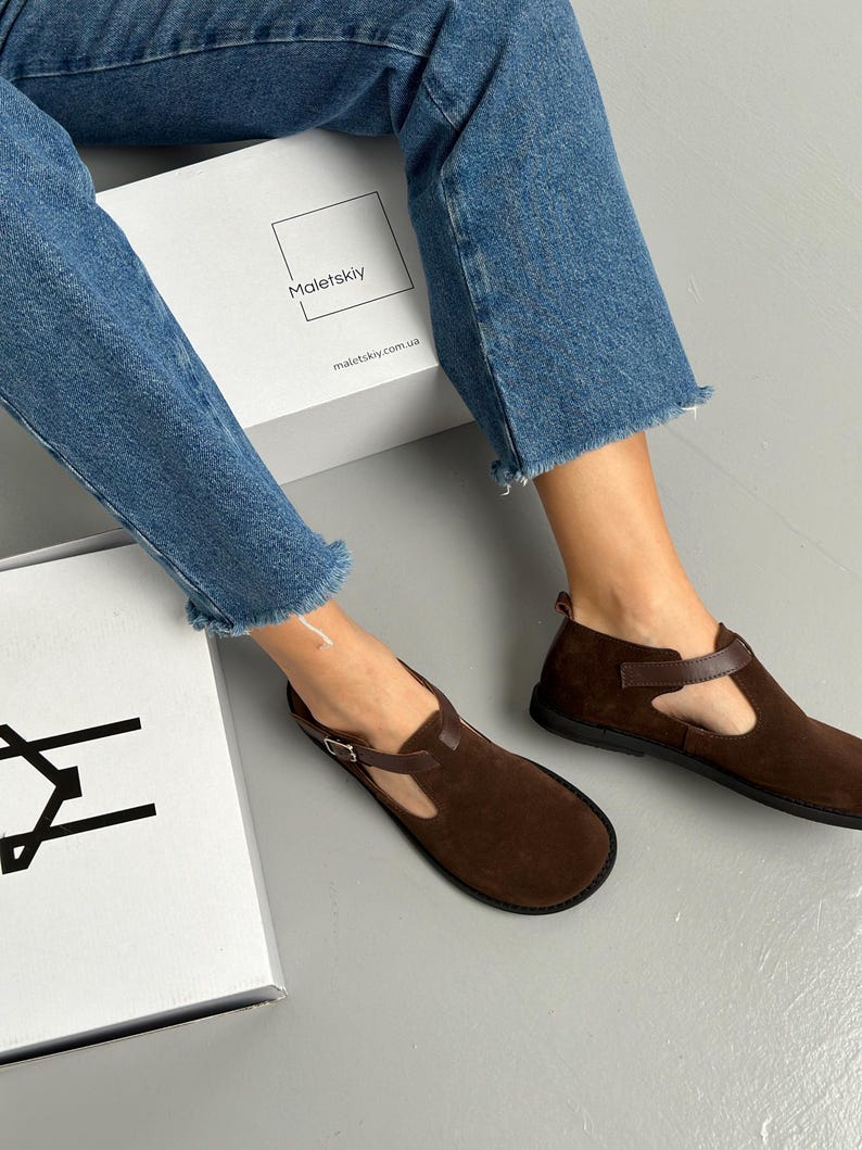 May include: Brown suede flats with a T-strap design and buckle closure. The shoes have a rounded toe and a dark brown leather strap. The shoes are displayed with a white box that has the text "Maletskiy" and "maletskiy.com.ua".