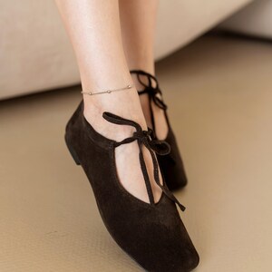 May include: Dark brown suede flats with a square toe and tie-up laces. The shoes are low-heeled and have a minimalist design. A silver anklet with small beads is also visible.