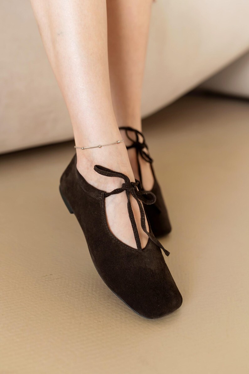 May include: Dark brown suede flats with a square toe and tie-up laces. The shoes are low-heeled and have a minimalist design. A silver anklet with small beads is also visible.