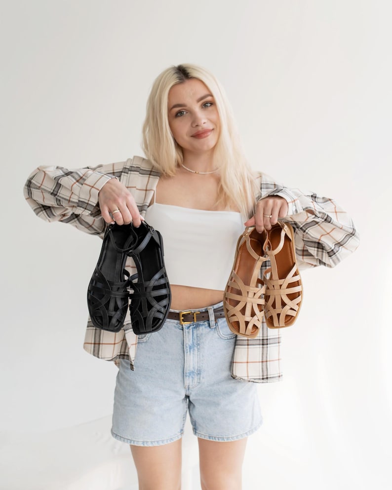 May include: A person holding two pairs of sandals. One pair is black, the other is tan. Both have a strappy design. The person is wearing a white top, denim shorts, and a plaid shirt.