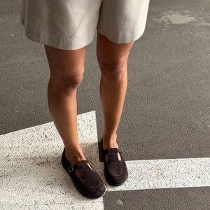 May include: A pair of dark brown suede Mary Jane style shoes with a buckle closure. The shoes have a low heel and are worn with light-colored shorts. The image is taken from a low angle, showing the shoes and the lower legs.