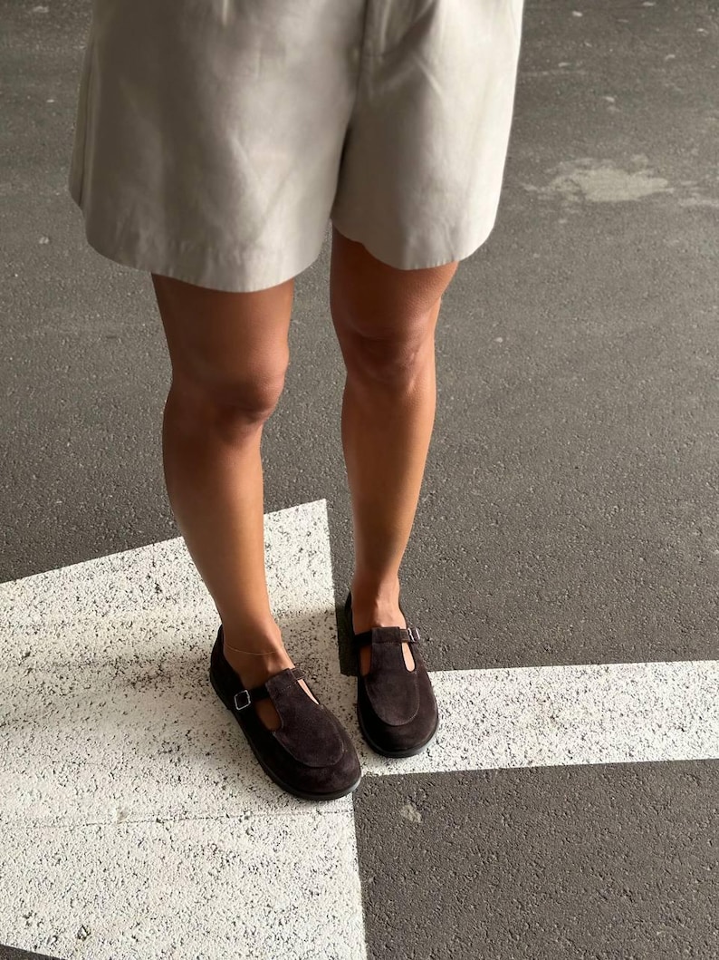 May include: A pair of dark brown suede Mary Jane style shoes with a buckle closure. The shoes have a low heel and are worn with light-colored shorts. The image is taken from a low angle, showing the shoes and the lower legs.