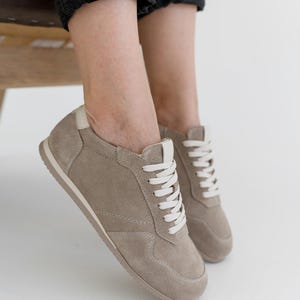Pu&ograve; includere: Sneakers in camoscio beige con lacci e suola bianchi. Le scarpe hanno un profilo basso e un design classico. Le sneakers sono mostrate con jeans neri. Le scarpe sono di colore neutro e sarebbero adatte all'uso quotidiano.