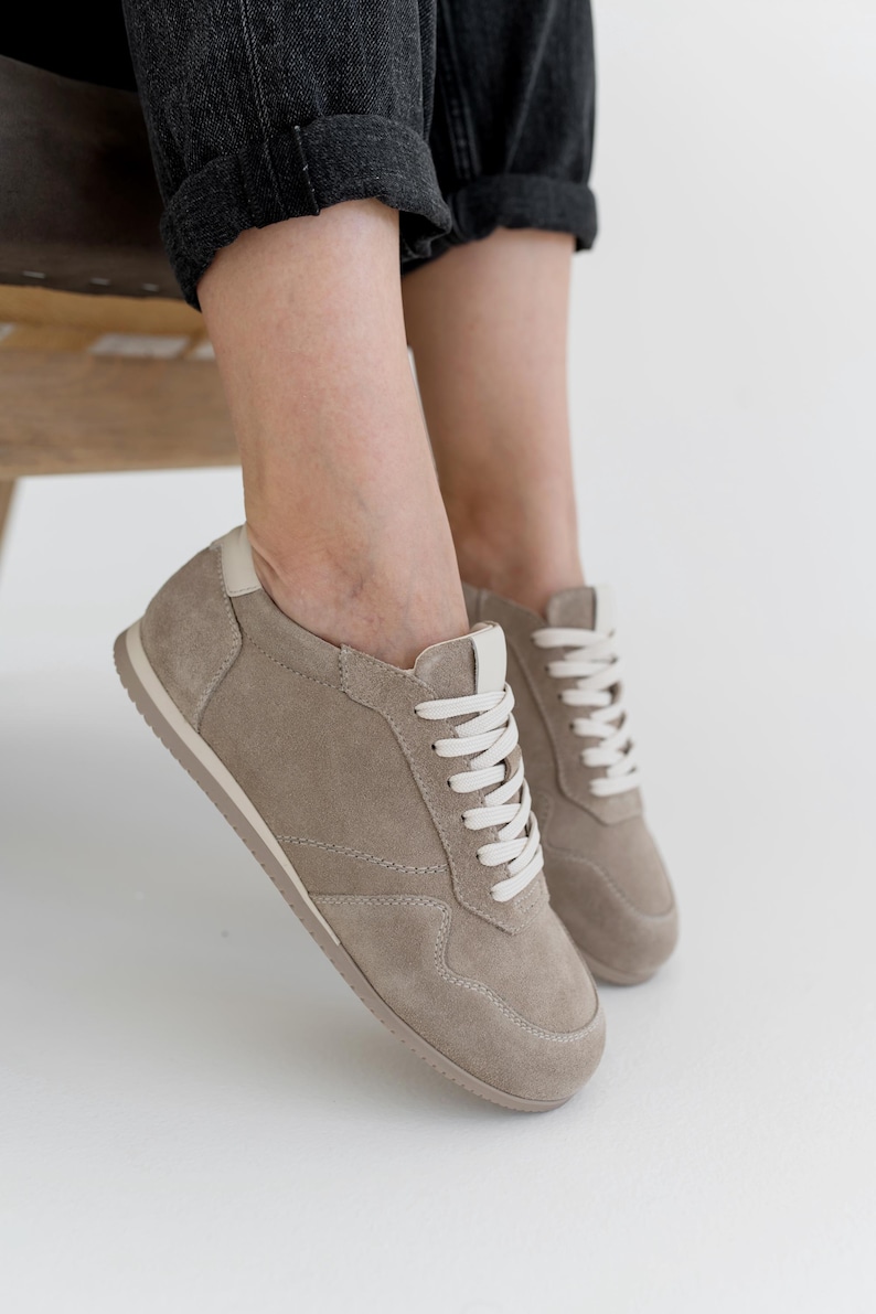 Pu&ograve; includere: Sneakers in camoscio beige con lacci e suola bianchi. Le scarpe hanno un profilo basso e un design classico. Le sneakers sono mostrate con jeans neri. Le scarpe sono di colore neutro e sarebbero adatte all'uso quotidiano.