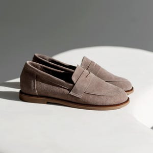 May include: A pair of taupe suede penny loafers with a light brown sole. The shoes are displayed on a white surface, with shadows adding depth. The classic design features a strap across the vamp.
