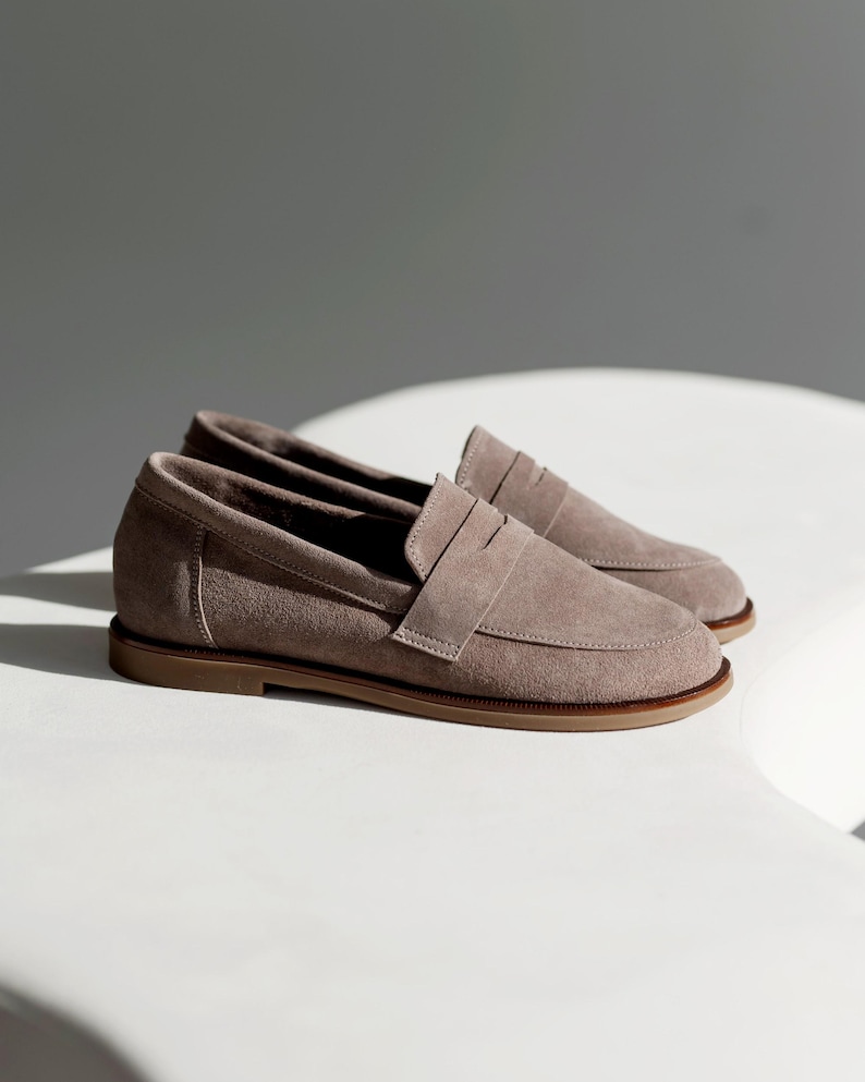 May include: A pair of taupe suede penny loafers with a light brown sole. The shoes are displayed on a white surface, with shadows adding depth. The classic design features a strap across the vamp.