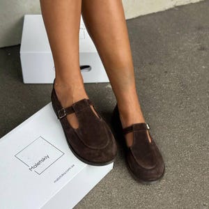 May include: Brown suede T-strap shoes with a buckle closure. The shoes are displayed on a white box with the brand name "Maletskiy" printed on it. The shoes are a dark brown color and have a flat sole.