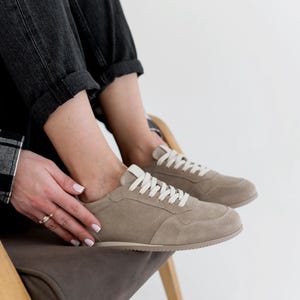Pu&ograve; includere: Sneakers in camoscio color cammello con lacci bianchi. Le scarpe basse hanno un design classico con suola cucita. Le sneakers sono indossate con jeans neri. Le scarpe sono di colore neutro e adatte all'uso quotidiano.