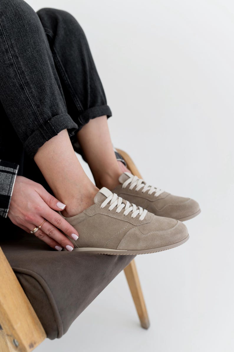 Pu&ograve; includere: Sneakers in camoscio color cammello con lacci bianchi. Le scarpe basse hanno un design classico con suola cucita. Le sneakers sono indossate con jeans neri. Le scarpe sono di colore neutro e adatte all'uso quotidiano.