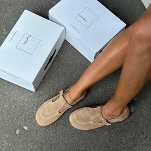 May include: Tan suede T-strap sandals with buckle closures. The sandals are displayed with two white boxes, each with the brand name "Maletskiy" printed on them. The sandals are on a gray surface.