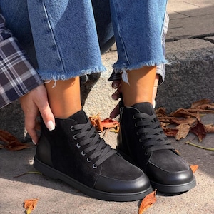 Könnte beinhalten: Ein Paar schwarze Stiefeletten aus einer Kombination von glattem Leder und Wildleder. Die Stiefel verfügen über schwarze Schnürsenkel, silberfarbene Ösen, eine abgerundete Zehenpartie, einen seitlichen Reißverschluss und eine flache Sohle.
