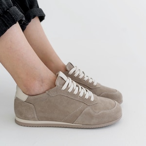 Pu&ograve; includere: Un paio di sneakers color talpa con lacci bianchi e un rinforzo bianco sul tallone. Le scarpe sono realizzate in materiale simile alla pelle scamosciata e presentano un design cucito. Le sneakers sono di colore neutro e adatte all'uso quotidiano.