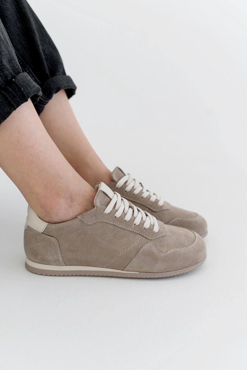 Pu&ograve; includere: Un paio di sneakers color talpa con lacci bianchi e un rinforzo bianco sul tallone. Le scarpe sono realizzate in materiale simile alla pelle scamosciata e presentano un design cucito. Le sneakers sono di colore neutro e adatte all'uso quotidiano.