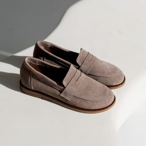 May include: A pair of taupe suede loafers with a penny strap detail. The shoes have a rounded toe and a brown sole. The loafers are displayed on a white surface, with shadows adding depth.