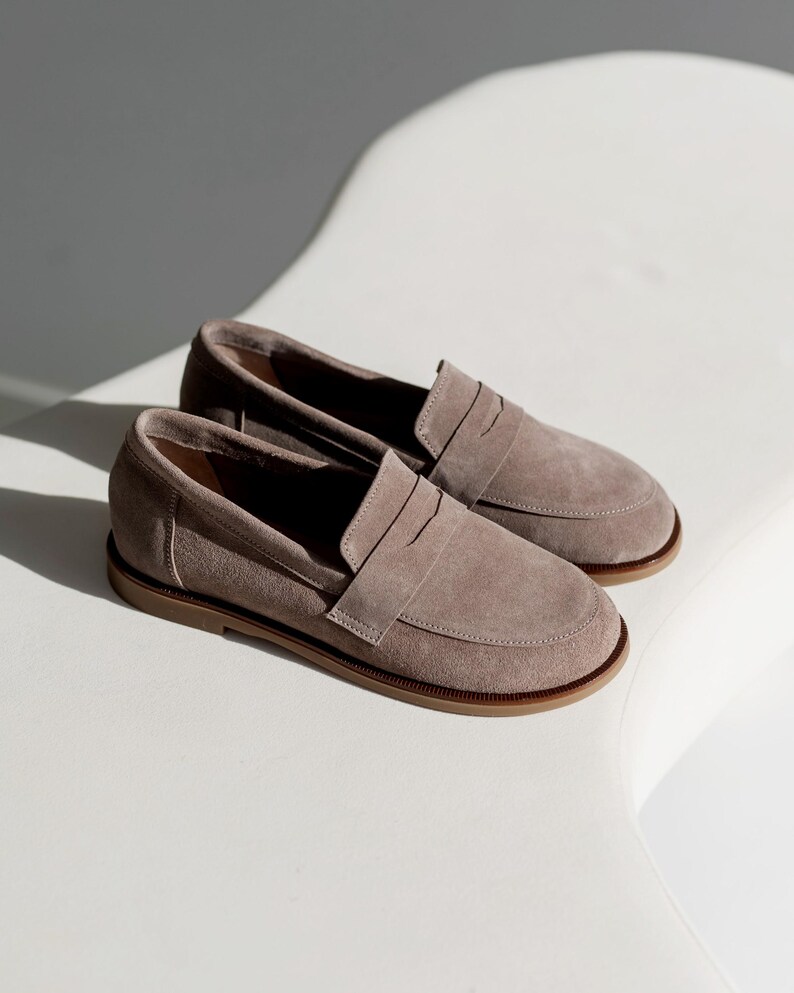 May include: A pair of taupe suede loafers with a penny strap detail. The shoes have a rounded toe and a brown sole. The loafers are displayed on a white surface, with shadows adding depth.