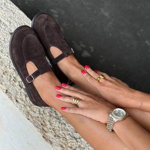 May include: A pair of brown suede Mary Jane style shoes with a buckle closure. The shoes are worn with gold rings and a silver watch. The person has red nail polish.