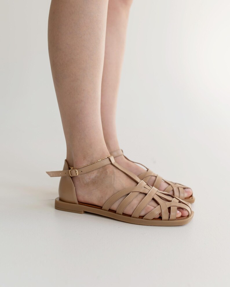 May include: Tan leather sandals with a cage-like design. The sandals have a flat sole and an adjustable ankle strap with a gold-colored buckle. The open-toe design features multiple straps across the foot.