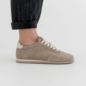 Pu&ograve; includere: Una sneaker in camoscio beige con lacci bianchi e un rinforzo sul tallone color crema. La scarpa ha un profilo basso e una suola in gomma. L'immagine mostra la scarpa di lato, con la caviglia e l'orlo arrotolato di un paio di jeans grigio scuro.