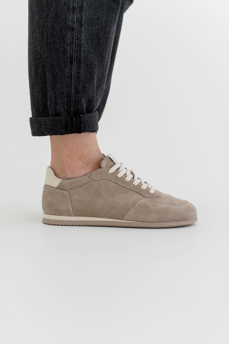 Pu&ograve; includere: Una sneaker in camoscio beige con lacci bianchi e un rinforzo sul tallone color crema. La scarpa ha un profilo basso e una suola in gomma. L'immagine mostra la scarpa di lato, con la caviglia e l'orlo arrotolato di un paio di jeans grigio scuro.