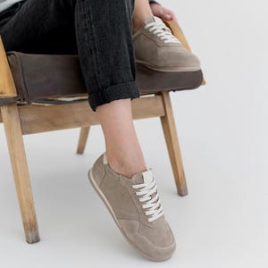 Pu&ograve; includere: Un paio di scarpe da ginnastica color talpa con lacci bianchi e un tallone bianco. Le scarpe sono realizzate in pelle scamosciata o materiale simile. Le scarpe da ginnastica sono mostrate su una persona seduta su una sedia di legno, con jeans neri.