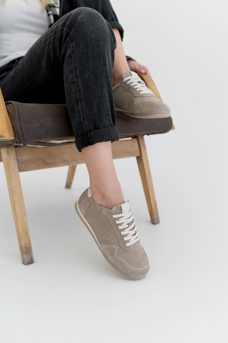 Pu&ograve; includere: Un paio di scarpe da ginnastica color talpa con lacci bianchi e un tallone bianco. Le scarpe sono realizzate in pelle scamosciata o materiale simile. Le scarpe da ginnastica sono mostrate su una persona seduta su una sedia di legno, con jeans neri.