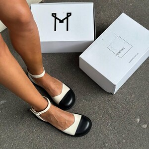 Puede incluir: Un par de zapatos planos bicolor con cuerpo color crema y puntera negra redondeada. Los zapatos tienen una tira fina al tobillo con hebilla met&aacute;lica. Hay dos cajas blancas, una con un logotipo de la letra M negra y la otra con Maletskiy.