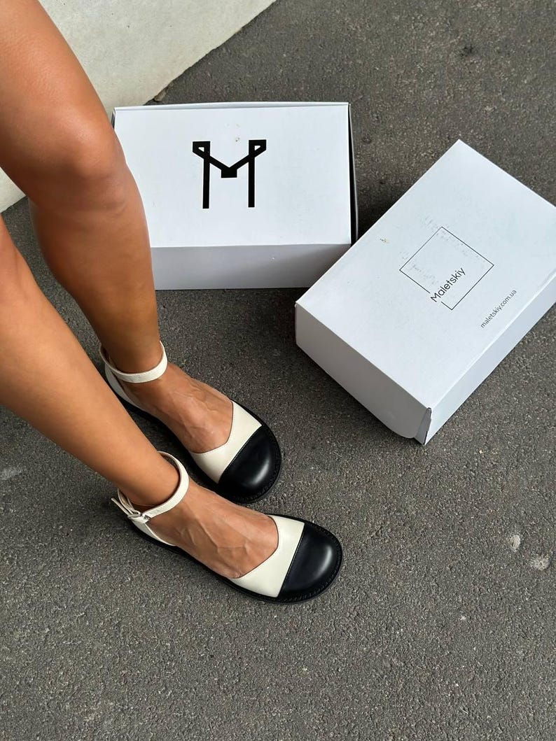 Puede incluir: Un par de zapatos planos bicolor con cuerpo color crema y puntera negra redondeada. Los zapatos tienen una tira fina al tobillo con hebilla met&aacute;lica. Hay dos cajas blancas, una con un logotipo de la letra M negra y la otra con Maletskiy.