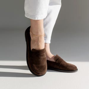 May include: A pair of dark brown suede loafers featuring a classic penny slot design across the vamp, a rounded toe, and a flat black sole. The shoes are worn with white trousers and a delicate gold chain anklet with small dangling charms.